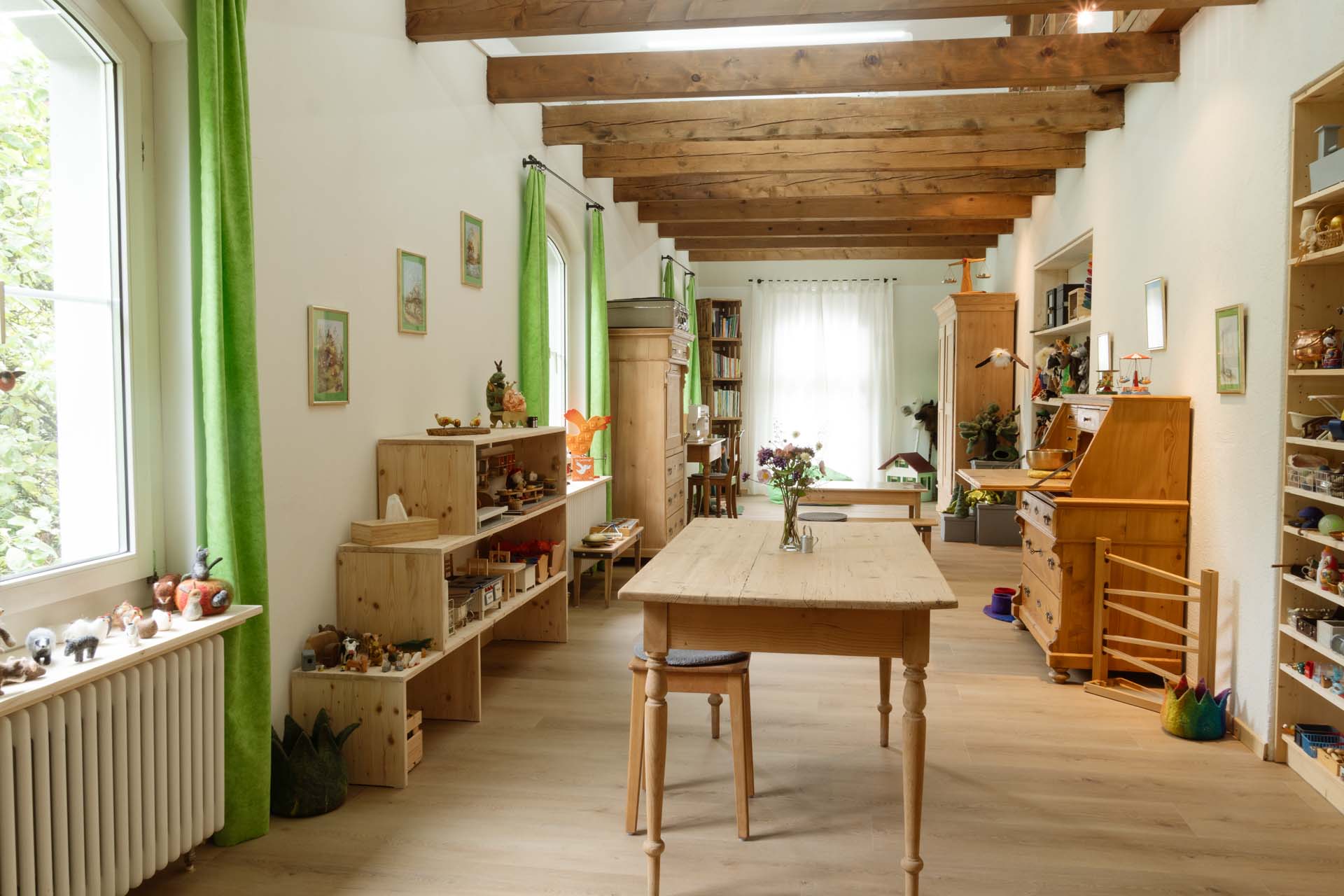 Heller, langer Praxisraum mit grünen Vorhängen, Holztisch in der Mitte und Holzmöbel auf der Seite.