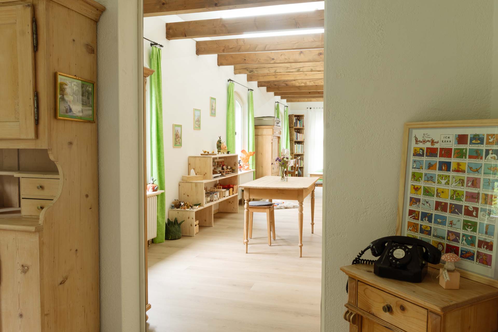 Blick durch die Tür in hellen, langen Praxisraum mit grünen Vorhängen, Holztisch in der Mitte und Holzmöbel auf der Seite.